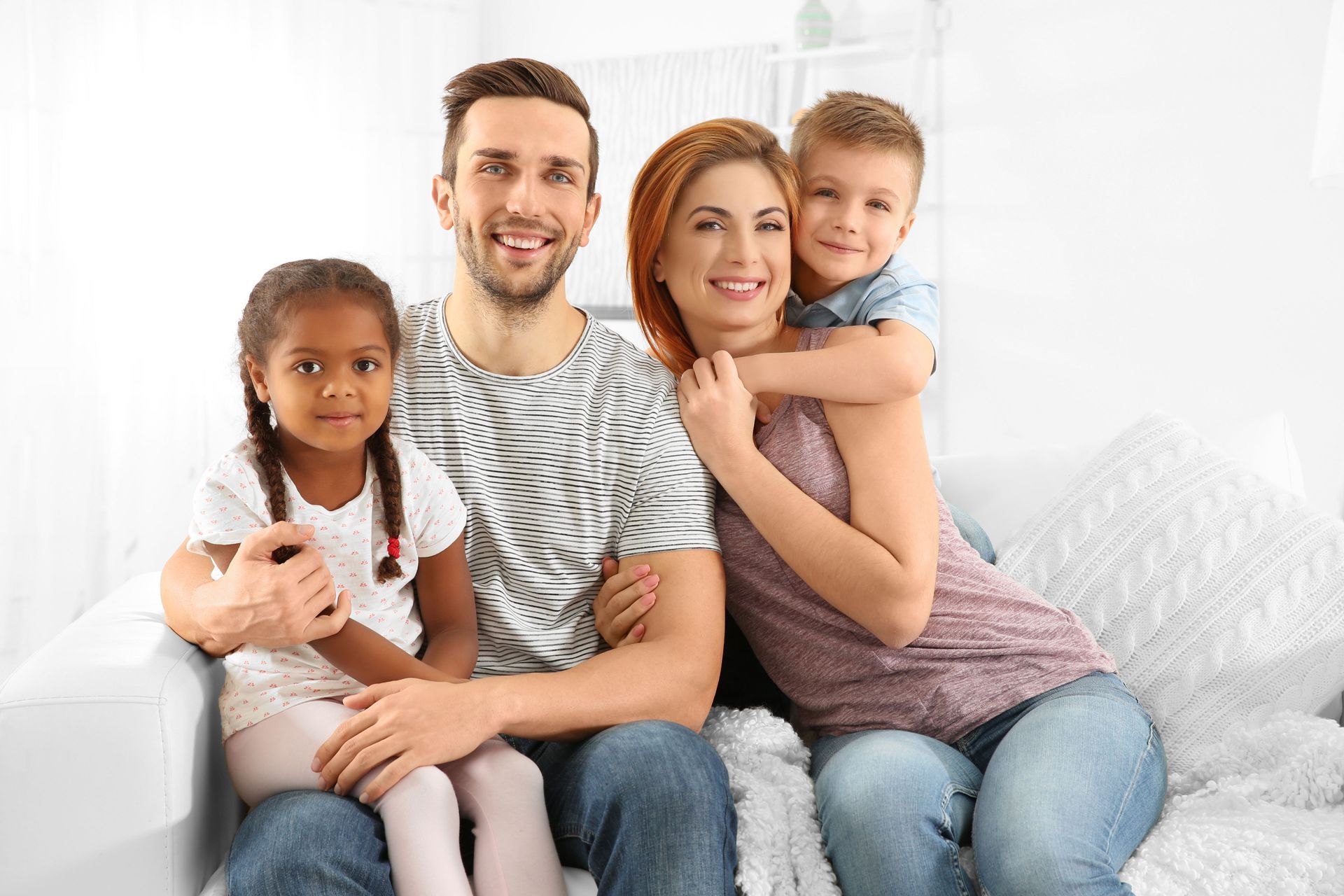 Family of four smiling on a white couch; two adults and two children.