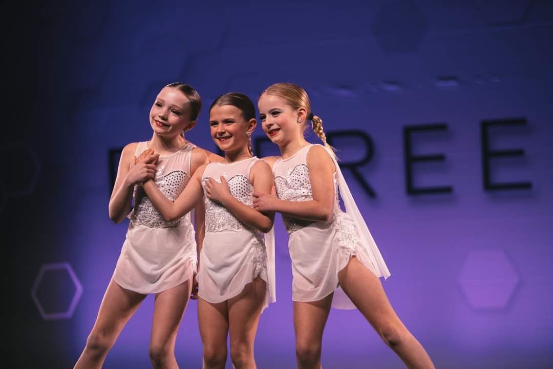Three young girls in white dresses are standing next to each other on a stage.