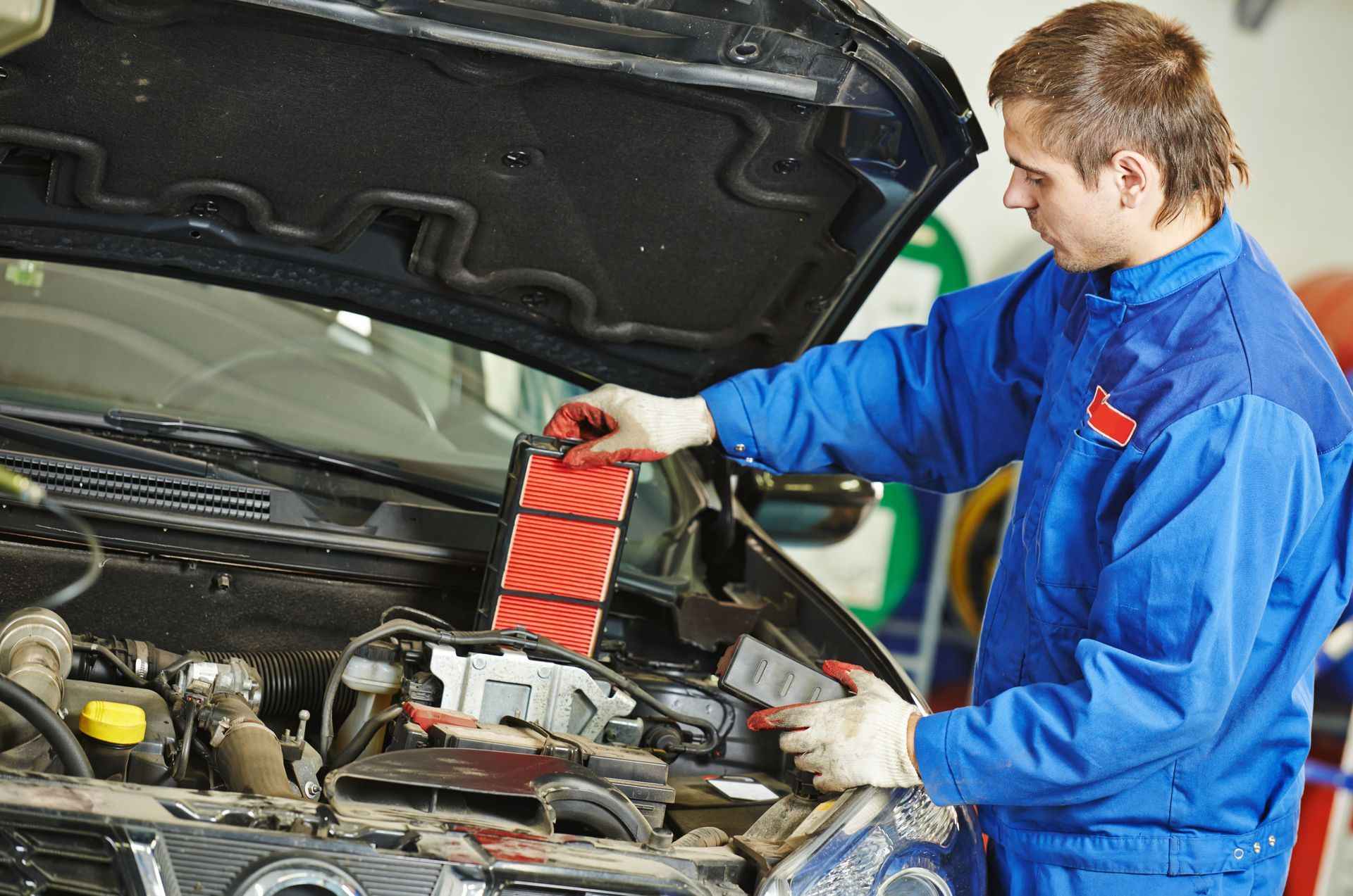 Mechanic in blue coveralls checks car engine with hood open and a red diagnostic device
