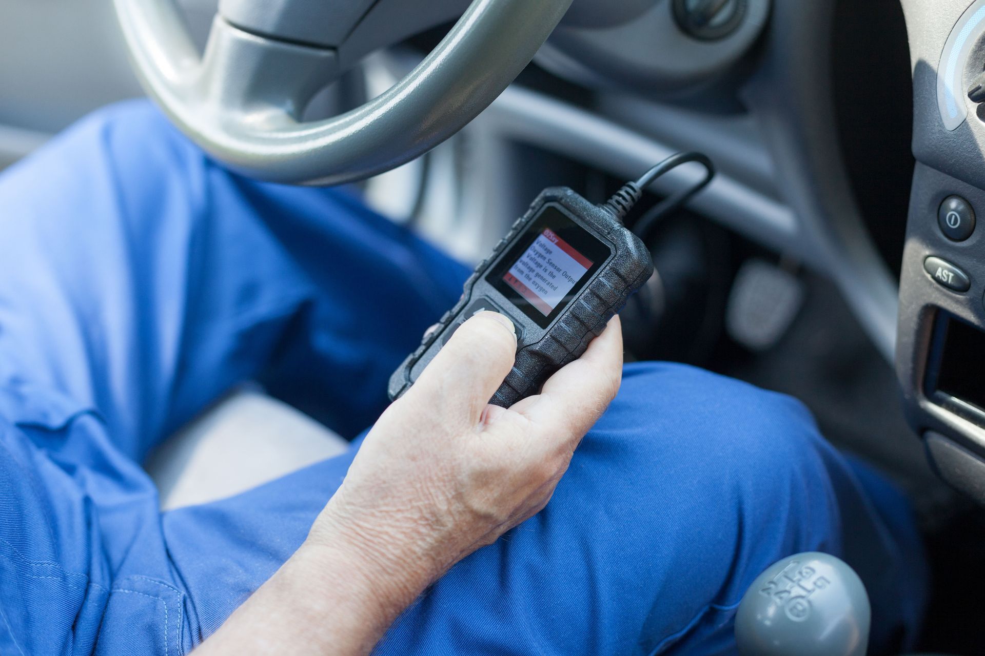 Driver holding a handheld diagnostic scanner inside a car, with blue pants and the steering wheel visible