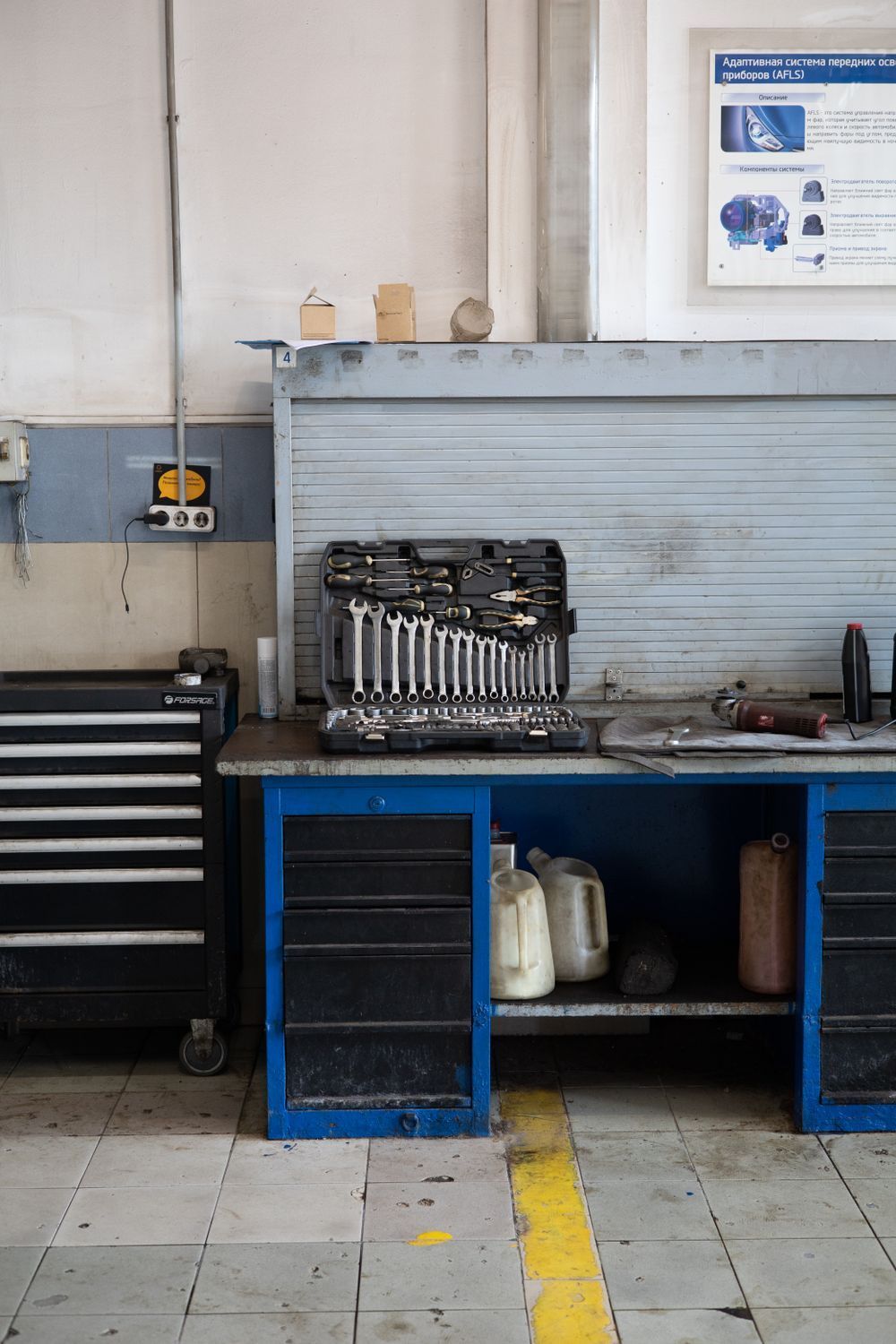 Workshop bench with stacked metal parts, blue cabinets, and a black tool chest against a tiled wall