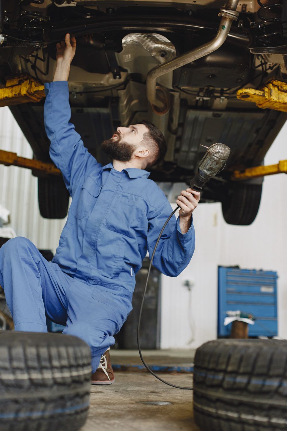 Mechanic in blue coveralls inspecting the underside of a car in a garage with tires on the floor
