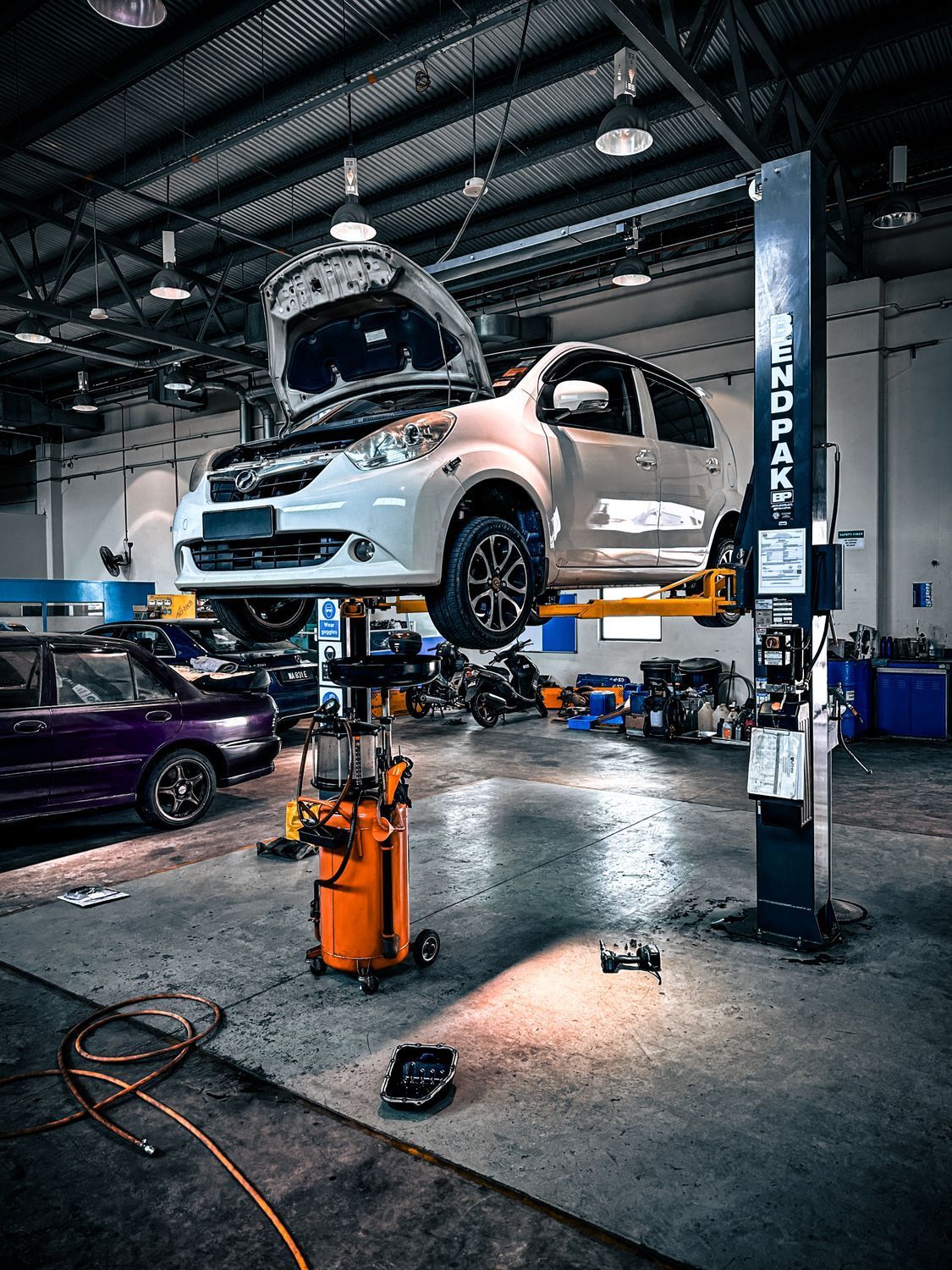 White car lifted in an auto repair shop, hood open, with tools and a mechanic nearby.