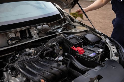 Open car hood showing engine bay and battery being worked on by a person nearby