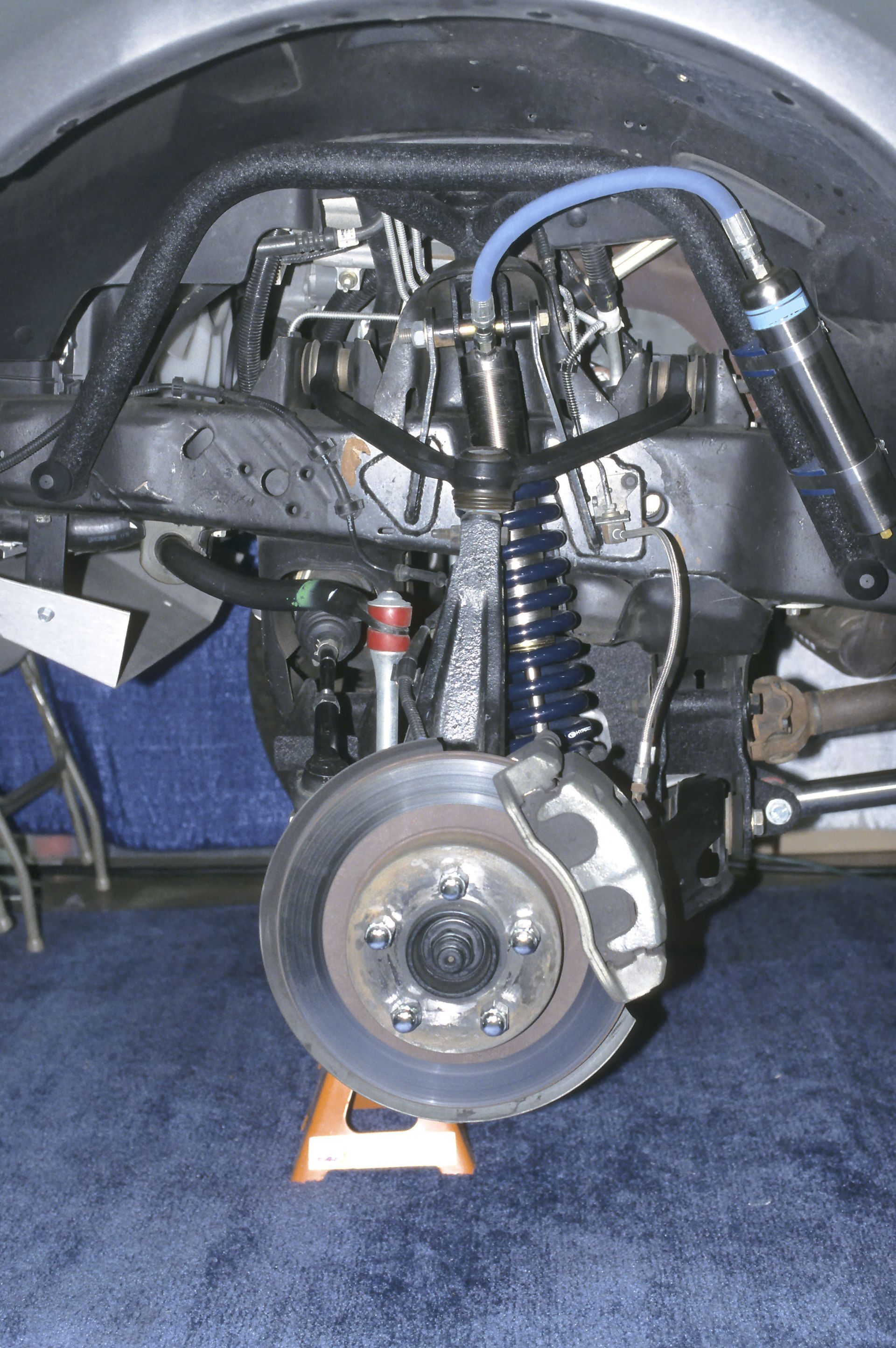 Car’s front suspension and brake assembly exposed in a garage, with wheel removed and blue hose visible