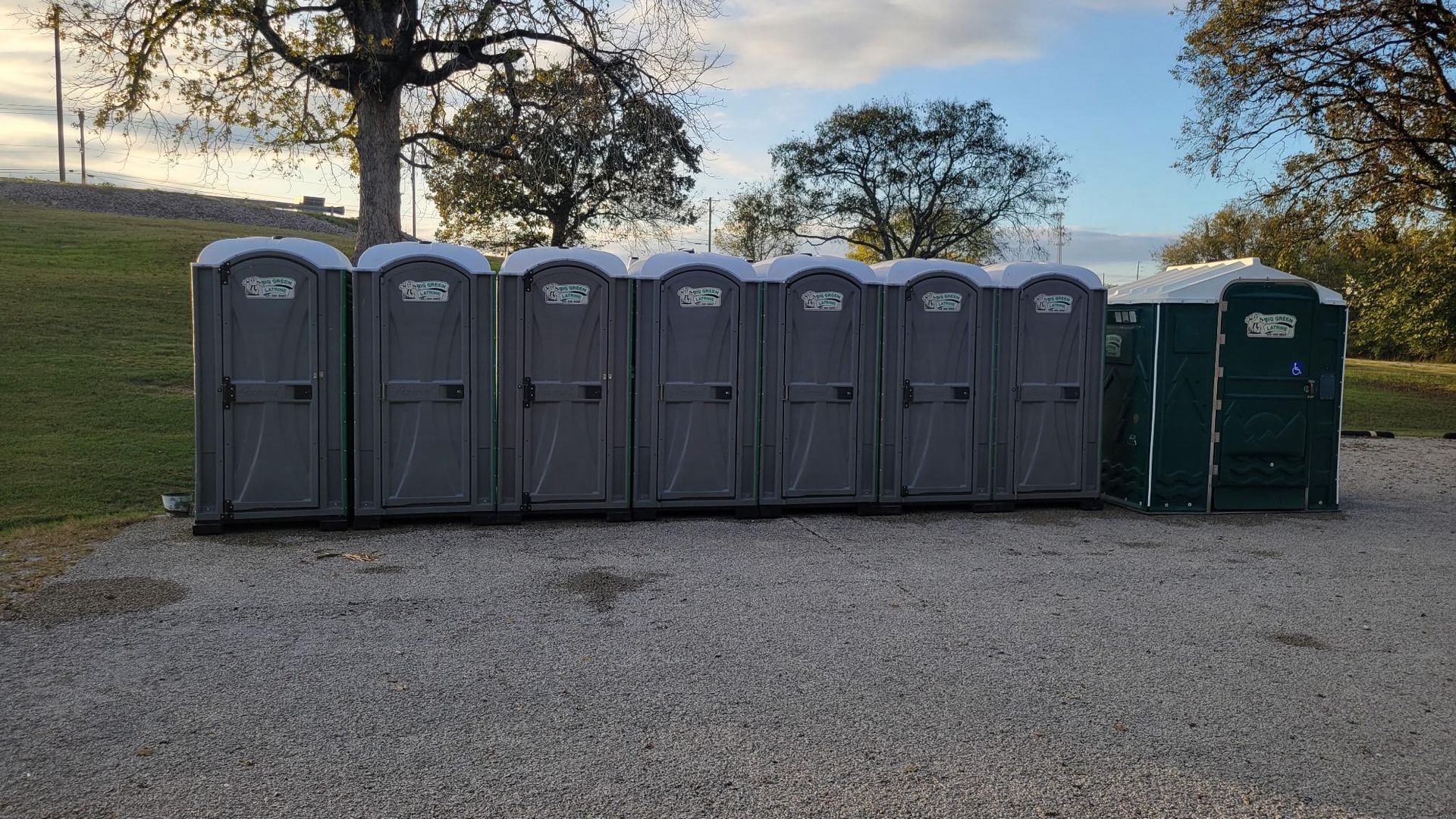 Row of gray and green portable toilets on gravel, with trees and sky in the background