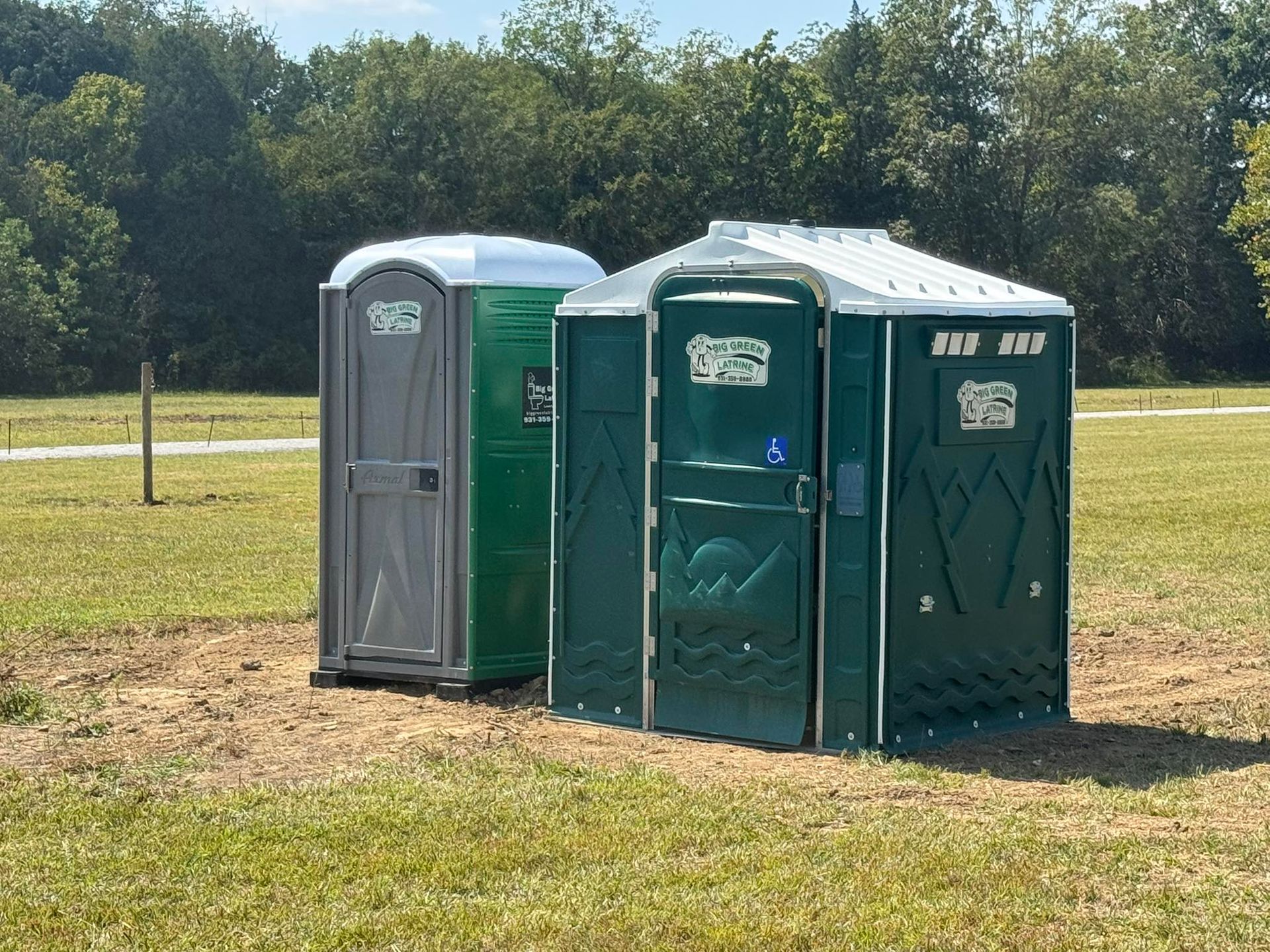 Two portable toilets, one gray and one green, standing on a grassy field near trees.