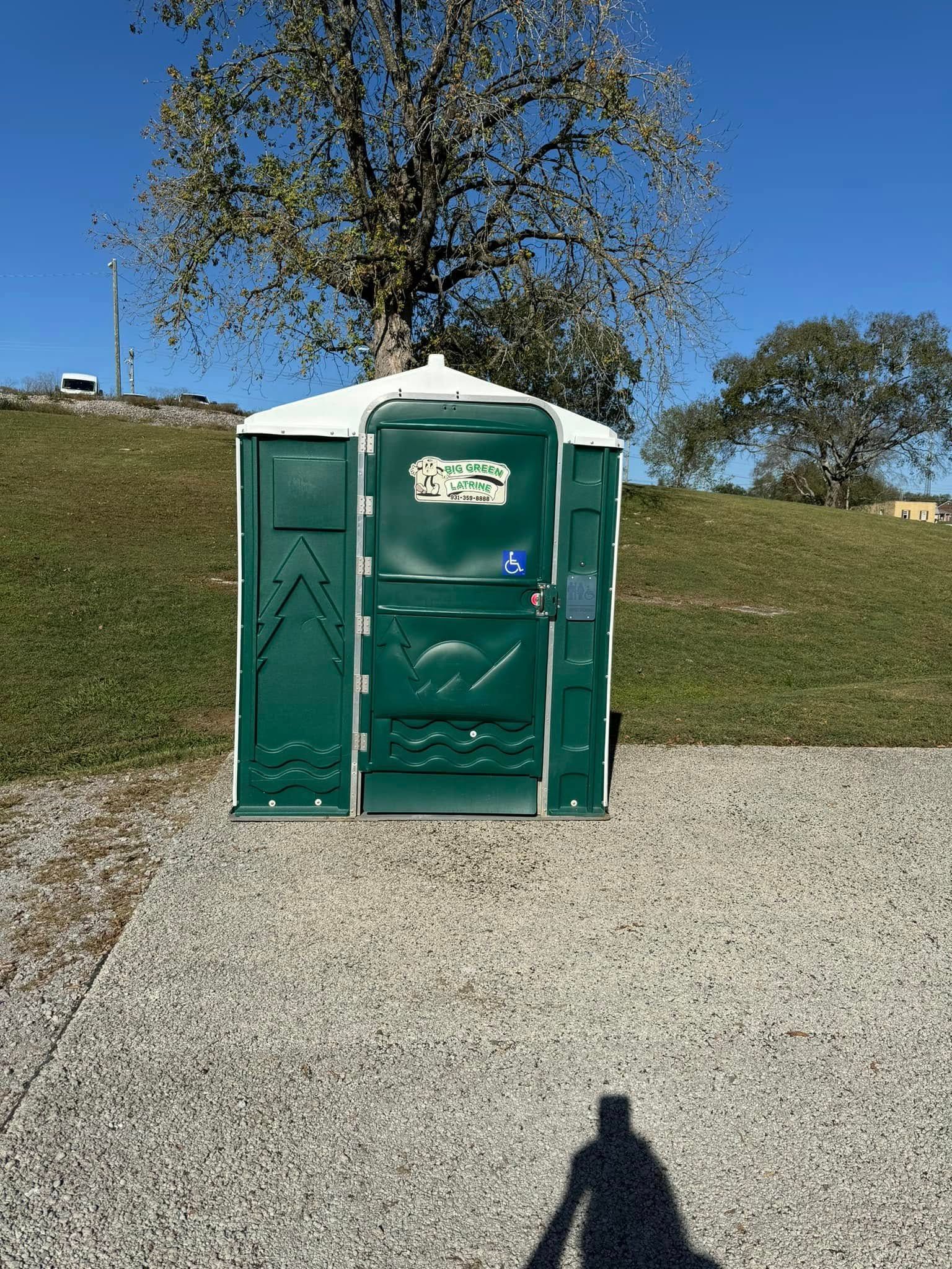 Green portable toilet on gravel beside a grassy hill and tree under a clear blue sky