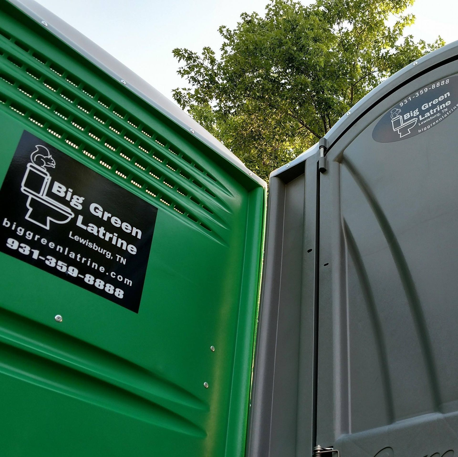Green portable toilet door with Big Green Lav line logo, beside a gray stall door outdoors under trees
