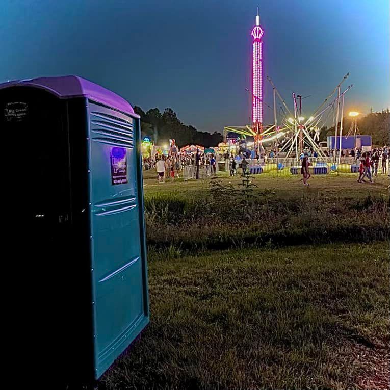 Portable toilet at a nighttime fair with neon rides and crowds in the background