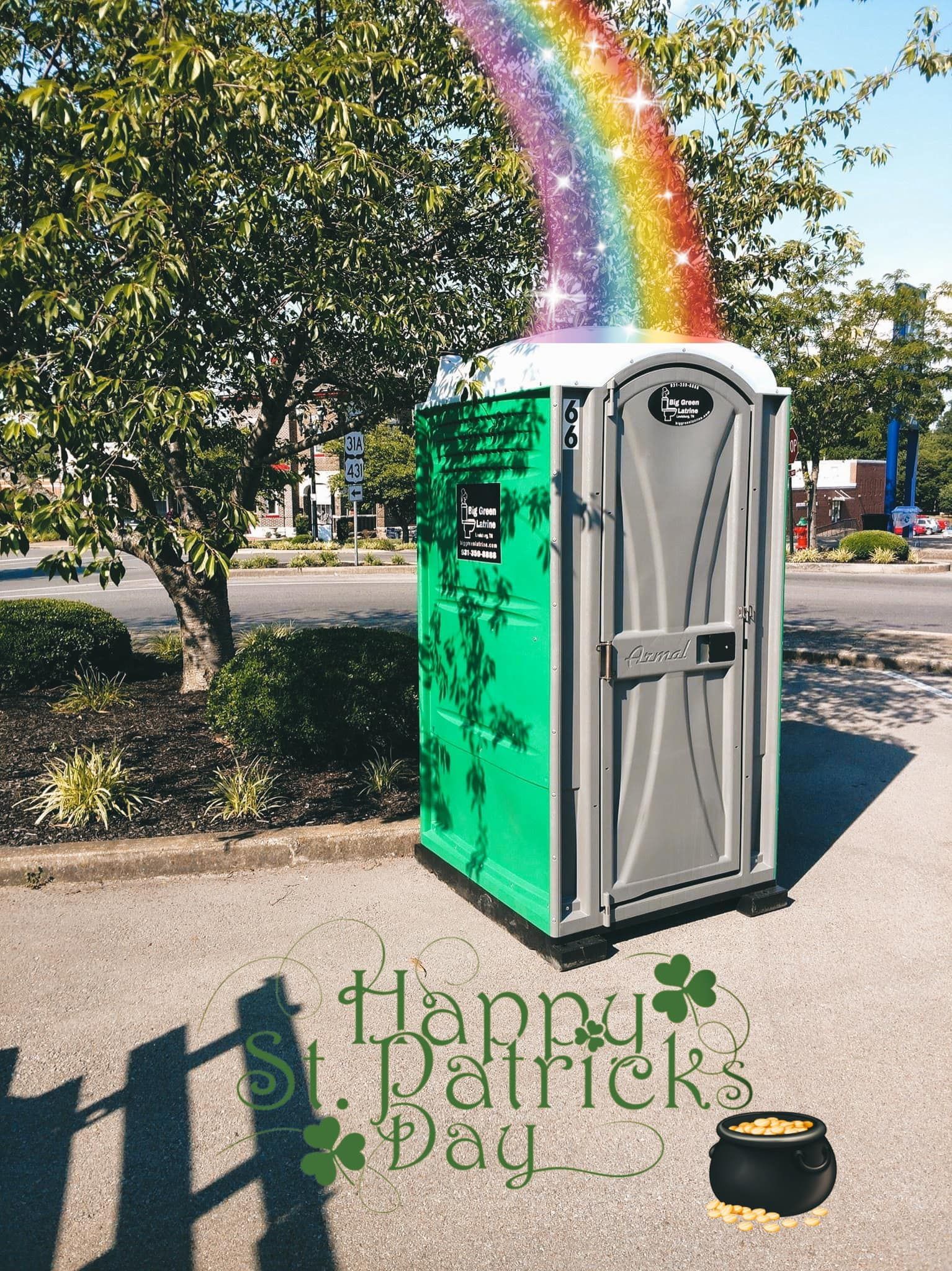 Portable toilet with a rainbow decoration beside a tree and clover-themed “Happy St. Patrick’s Day” sidewalk art.