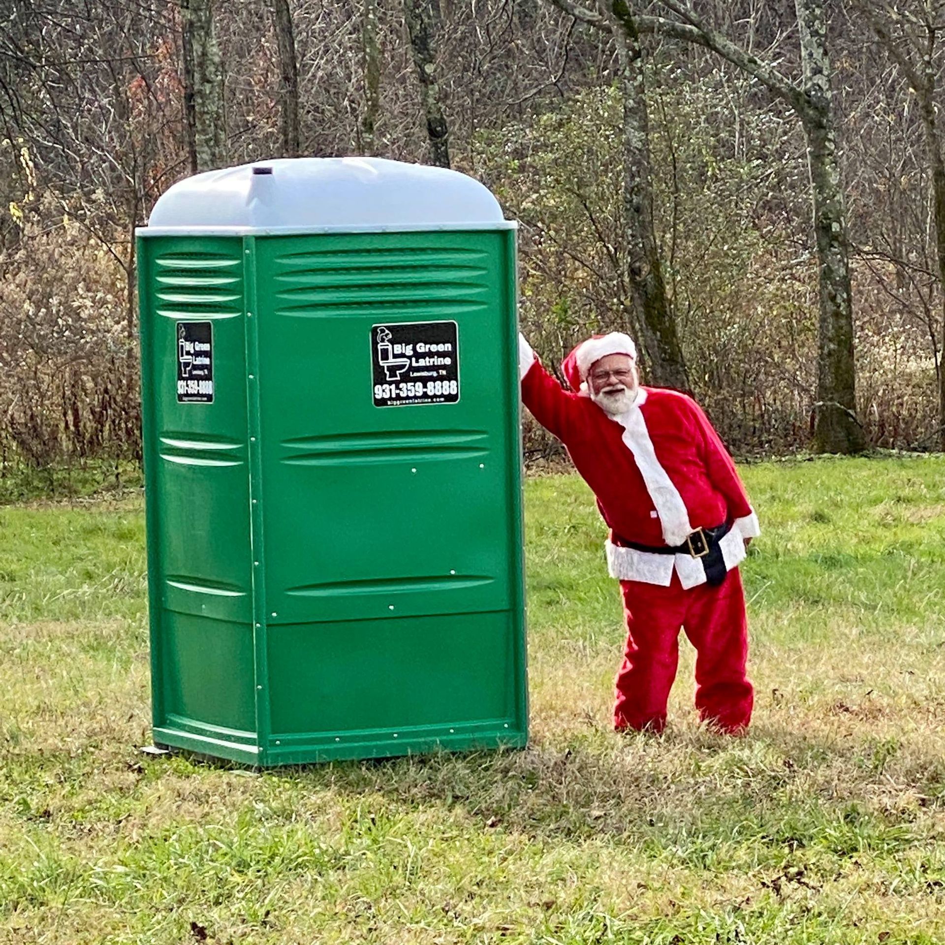 Santa in red suit posing beside a green portable toilet outdoors