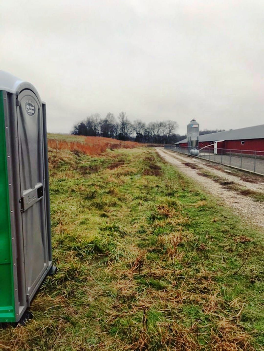 Grassy farm path beside a portable toilet, with red barns and a silo under a cloudy sky.