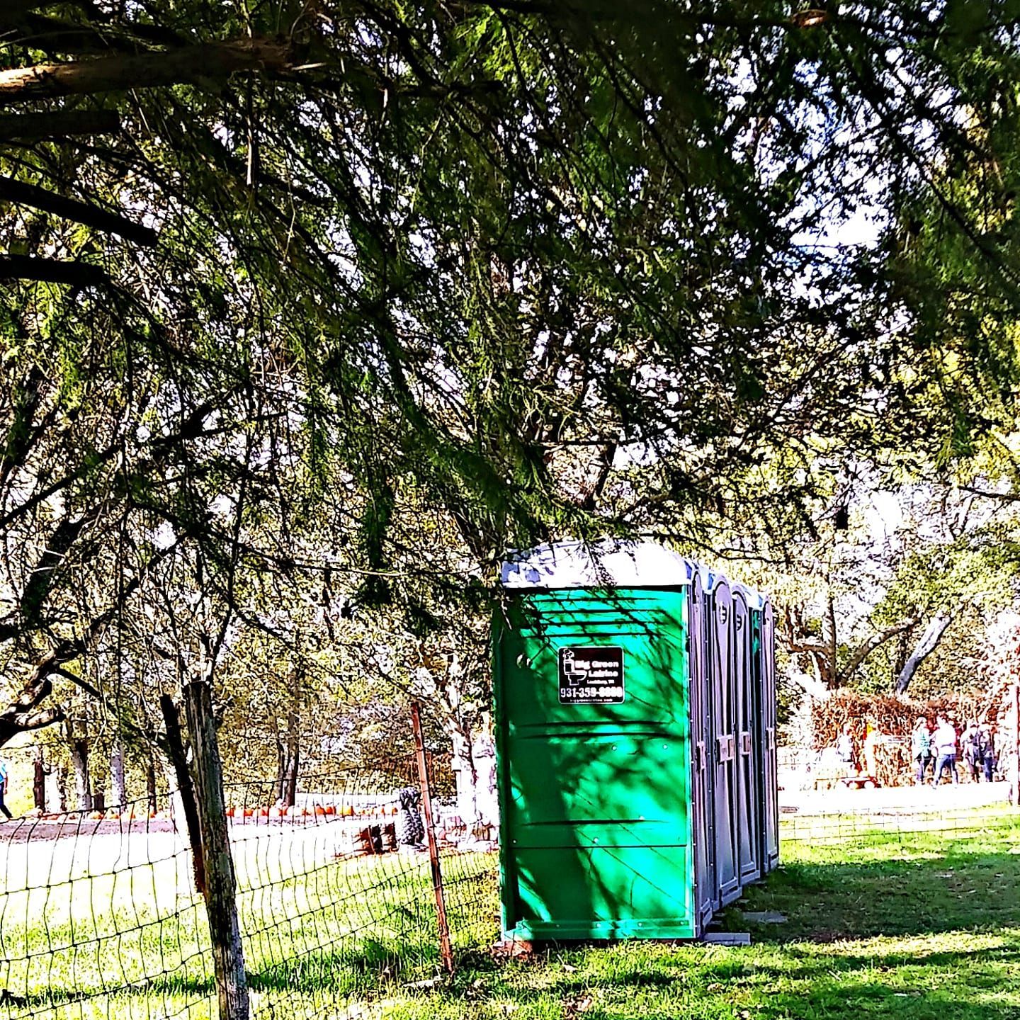 Green portable toilet in a shaded park beside a fence and trees