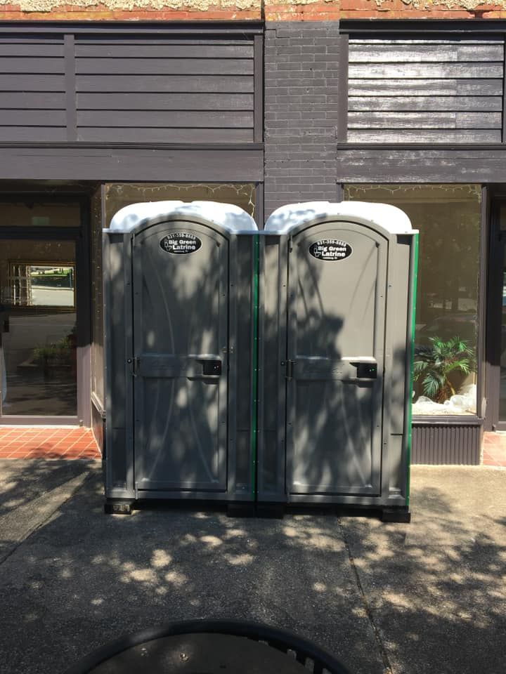 Two portable toilets beside a building, casting shadows on a sunny sidewalk.