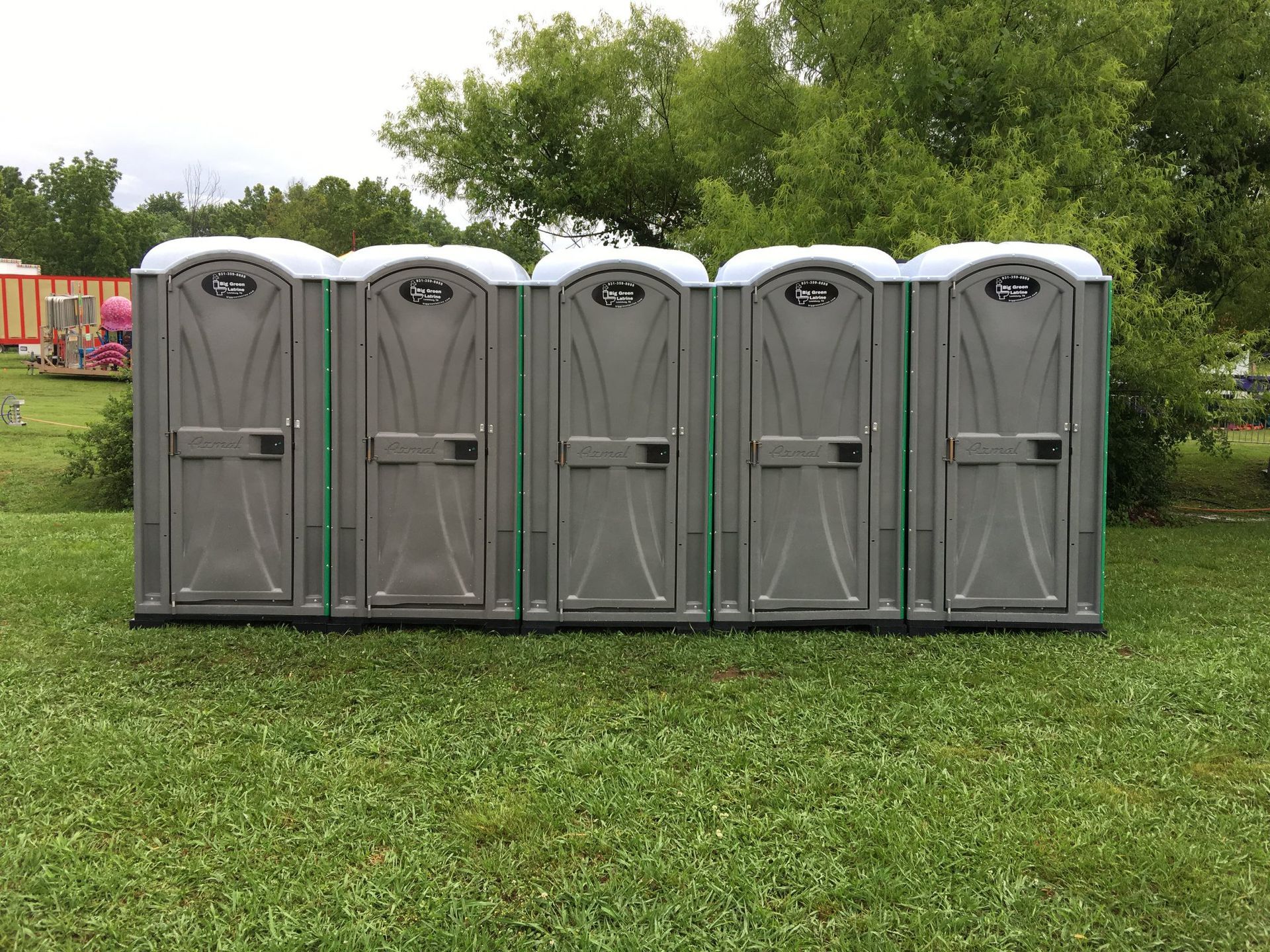 Row of gray portable toilets on a grassy field with trees in the background