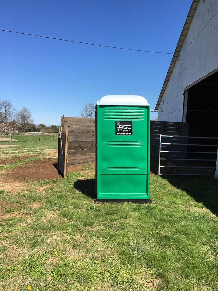 Green portable toilet beside a barn on a grassy farmyard under a clear blue sky