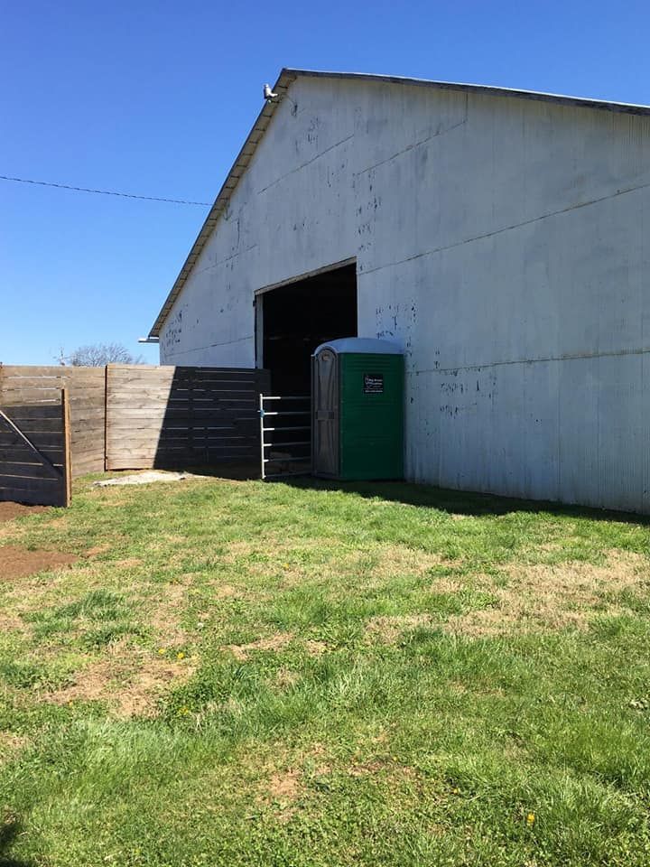 White barn-like building with open doorway and green portable toilet beside a grassy yard and fence