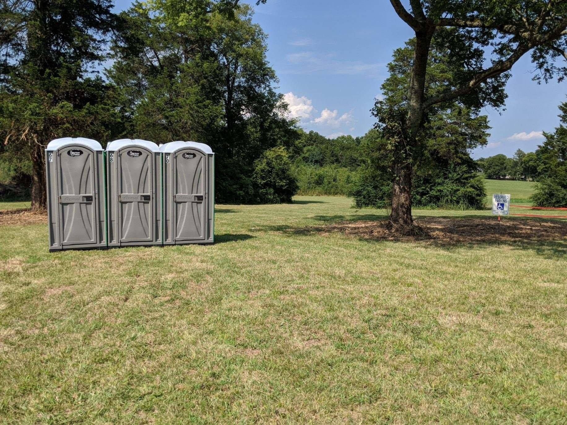 Three portable toilets in a grassy park with trees and a blue sky.