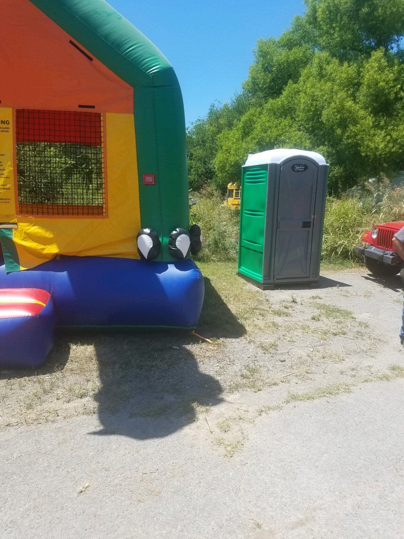 Colorful inflatable bounce house beside a green portable toilet on a sunny gravel lot.