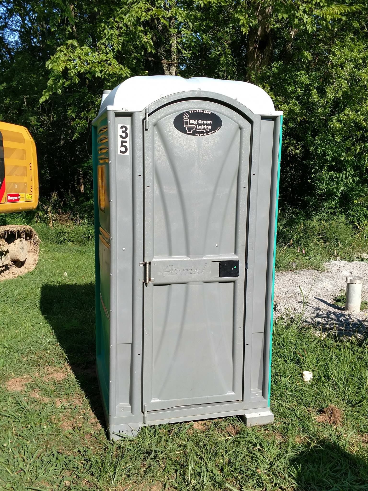 Portable gray portable toilet outdoors on grass near gravel and trees