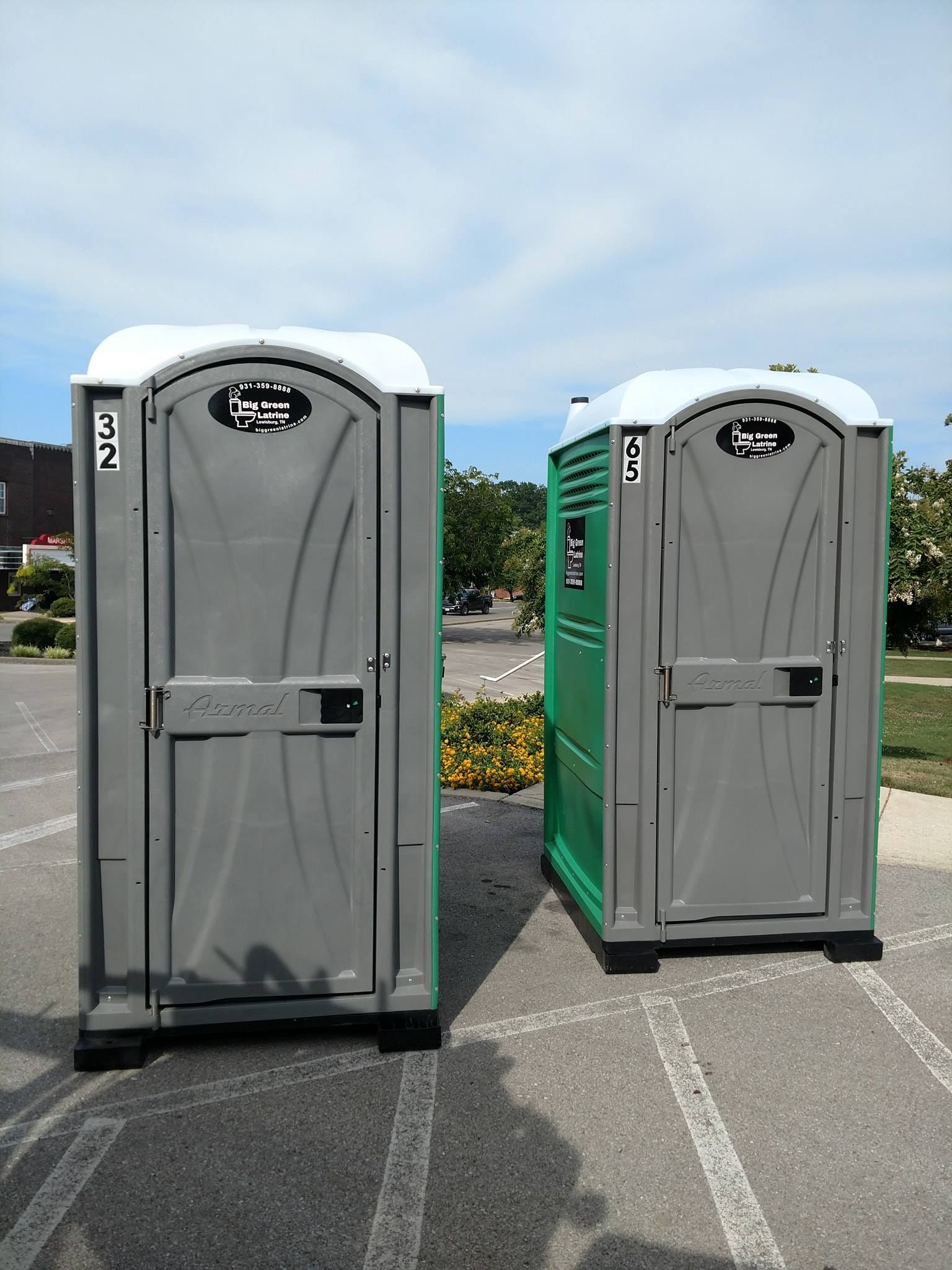 Two portable toilets, one gray and one green, standing in a parking lot under a blue sky.