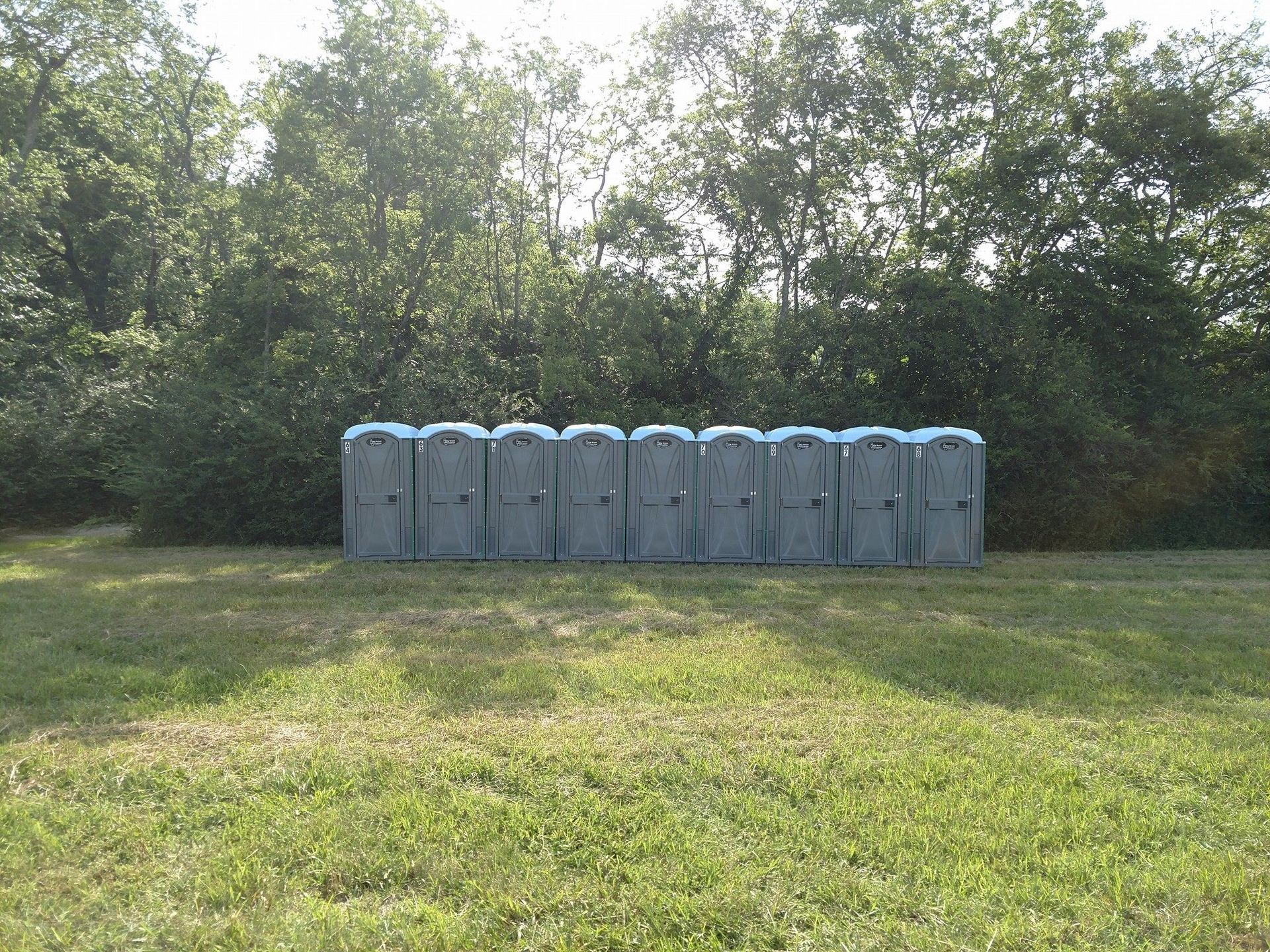 Row of gray portable toilets on a grassy field with trees in the background