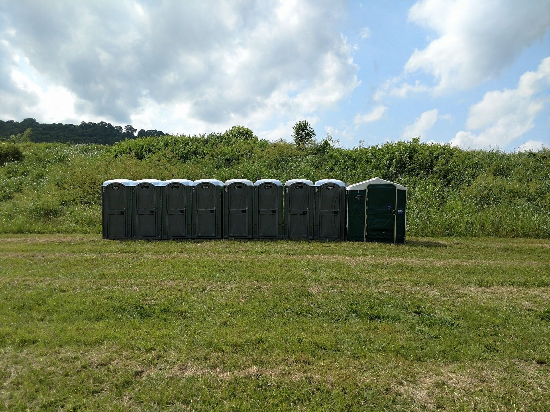Row of dark portable toilets beside a grassy field under a cloudy sky