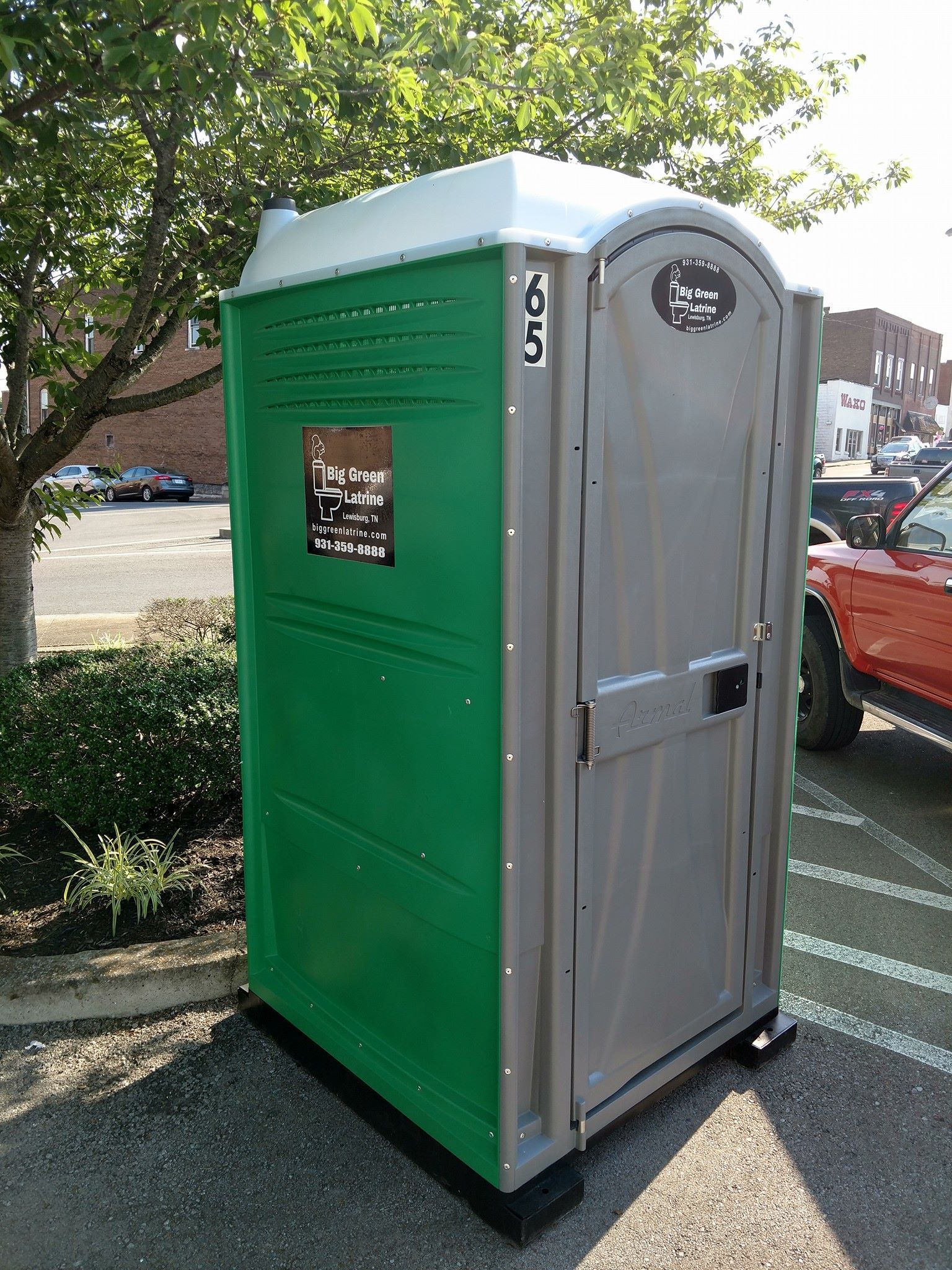 Portable green-and-gray portable toilet parked outdoors near trees and cars.