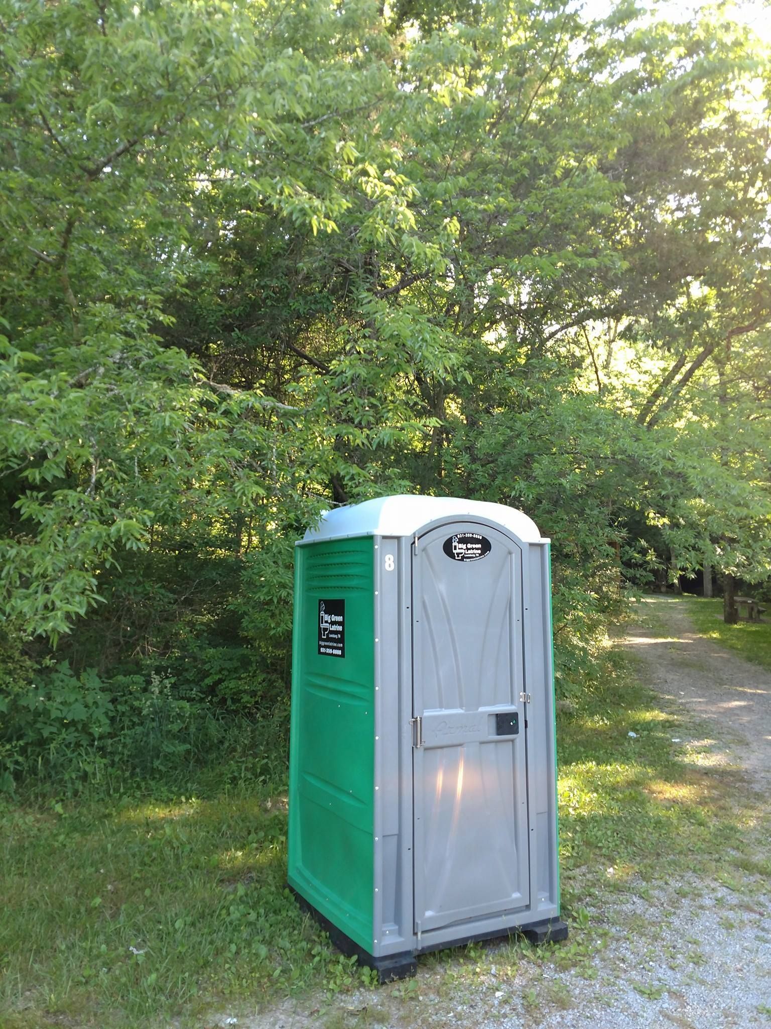 Green and gray portable toilet beside a wooded trail in a park
