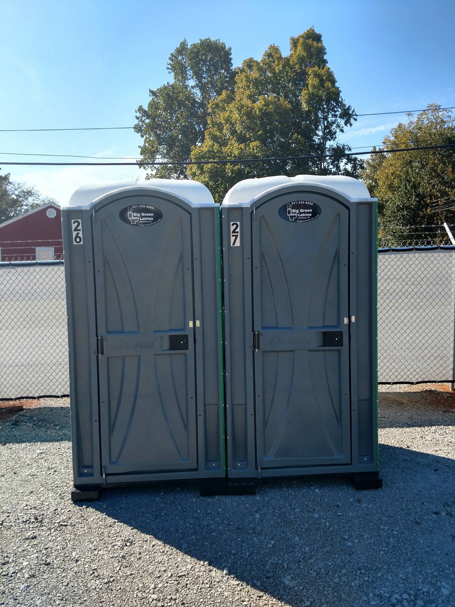 Two gray portable toilets on gravel in front of a fence and trees under a blue sky