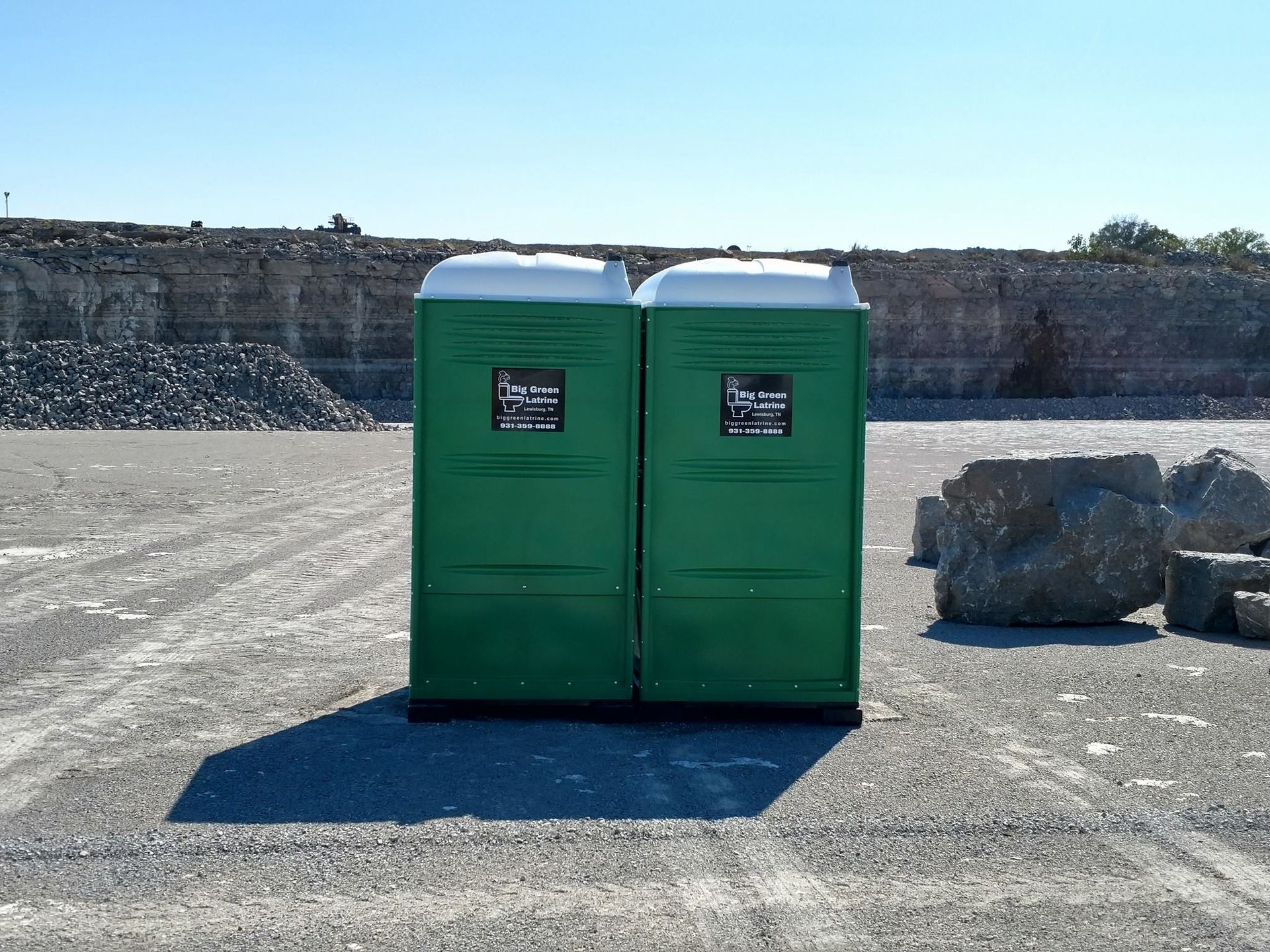 Two green portable toilets on a gravel lot with a stone wall in the background