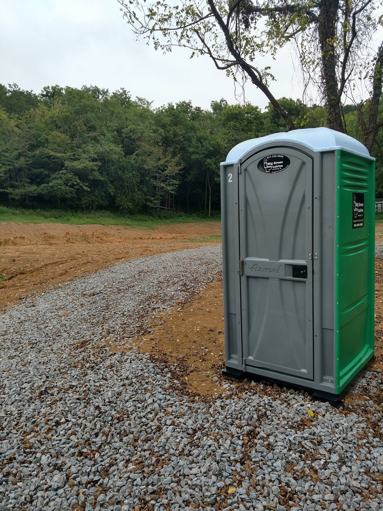 Portable toilet beside a gravel path in a muddy, wooded outdoor area