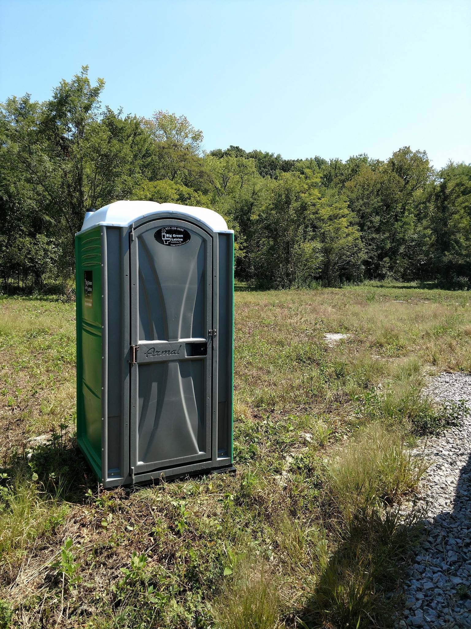 Portable toilet beside a grassy roadside with trees in the background