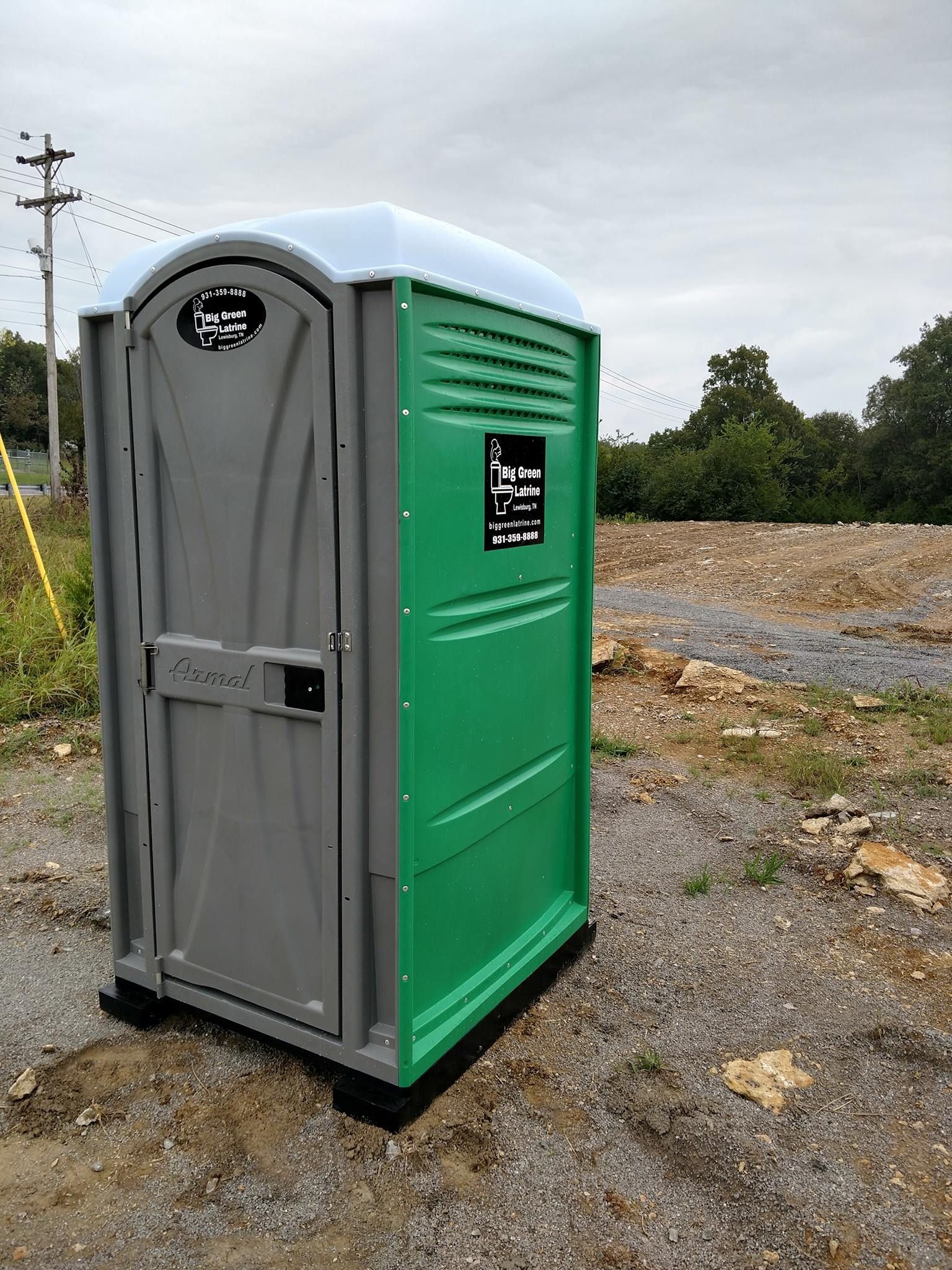 Portable toilet on gravel lot, with green side panels and gray door, outdoors near utility poles.