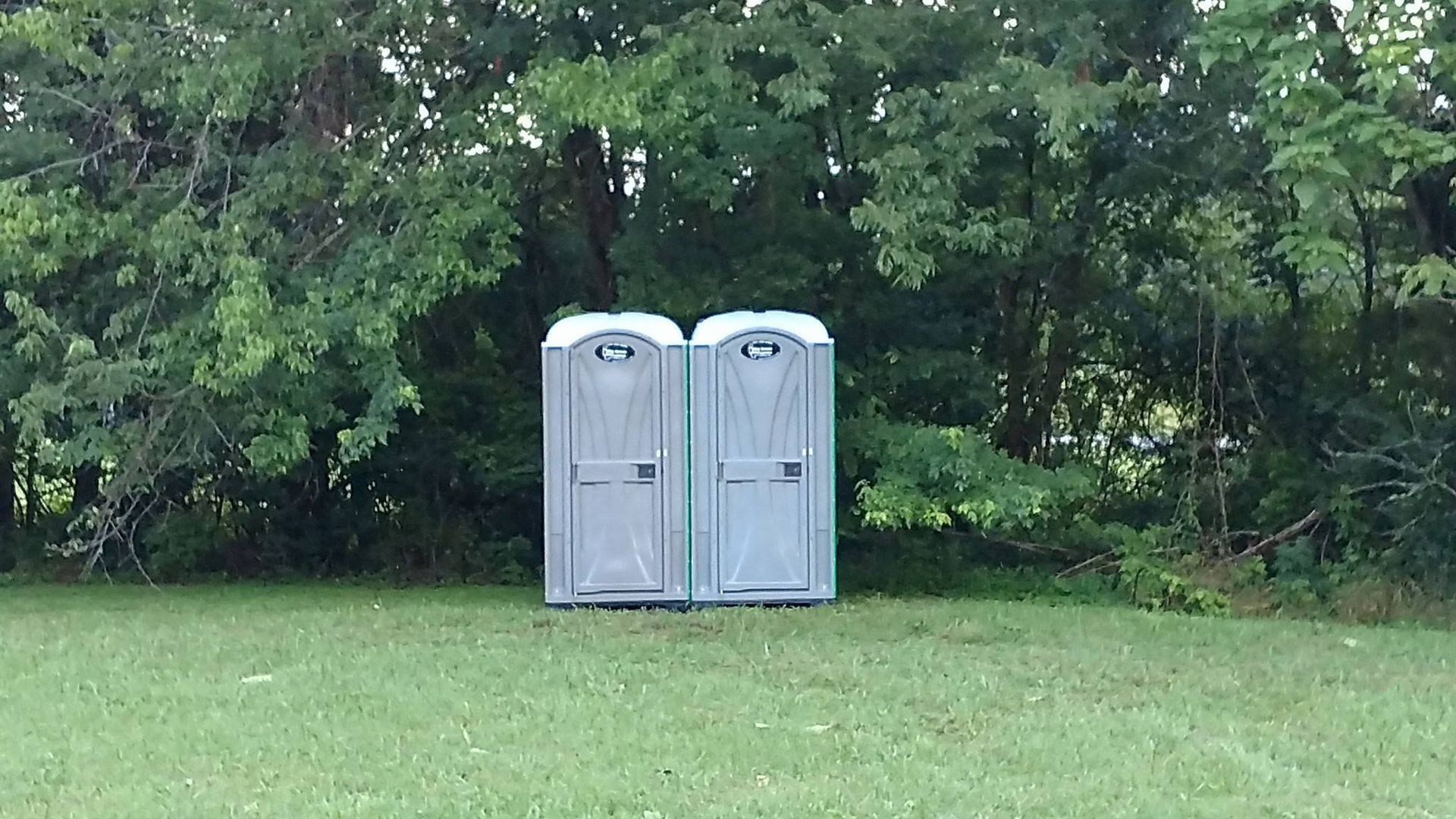 Two gray portable toilets on a grassy lawn in front of dense green trees.