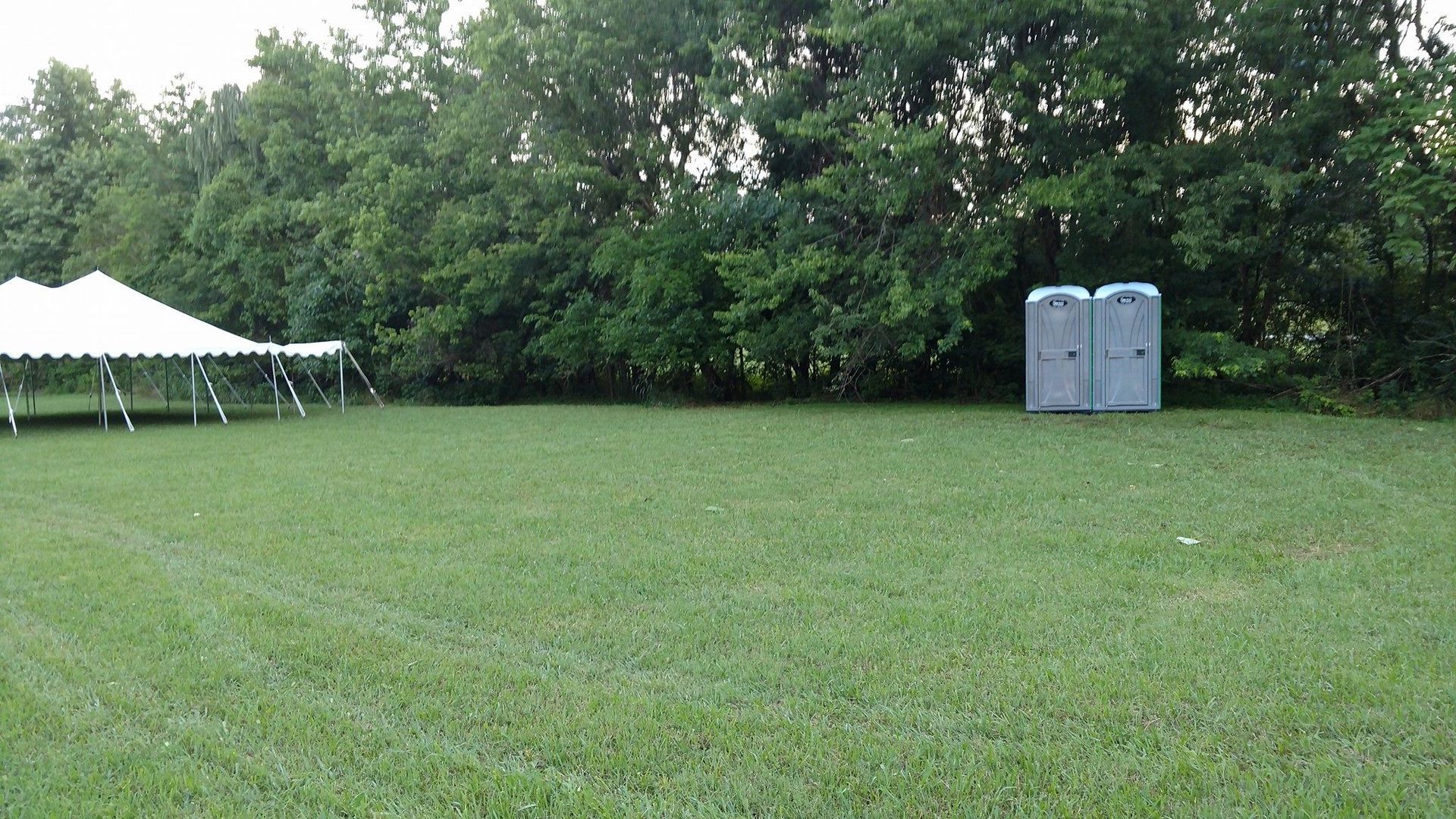 Grassy field with white tents on the left and two blue portable toilets near trees on the right.