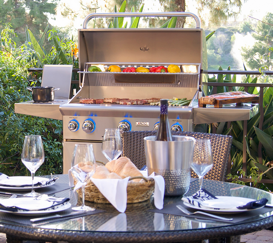 Outdoor grilling setup with food cooking. Table set with glasses, wine, and basket of bread.
