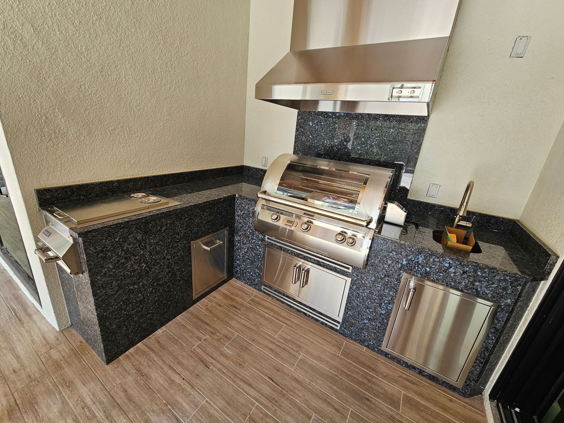 Outdoor kitchen with stainless steel grill, sink, and cabinets on granite countertops.