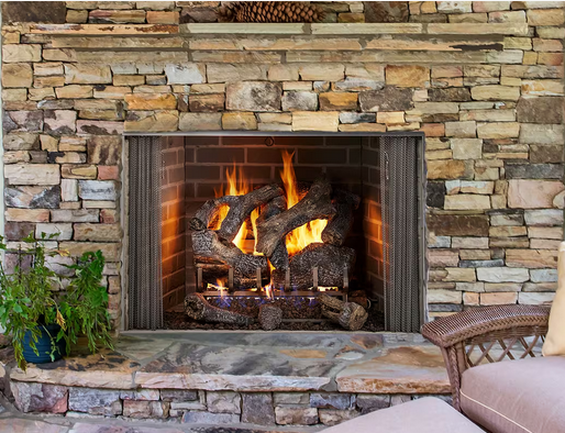 Fireplace with burning logs, inside a stone-covered hearth and surround. A potted plant and chair are visible.