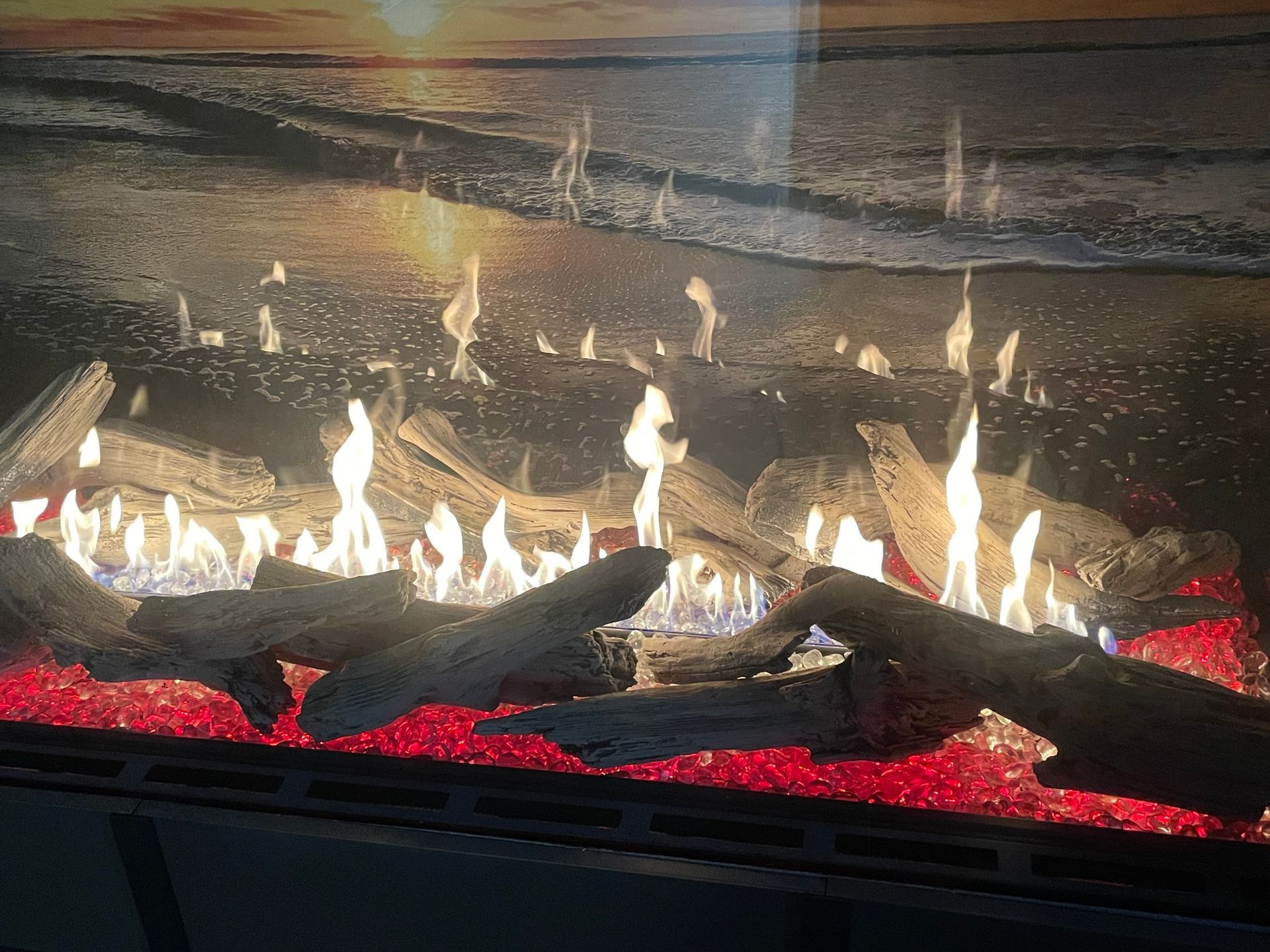 Fireplace with glowing red embers, driftwood, and flames against a beach scene backdrop.