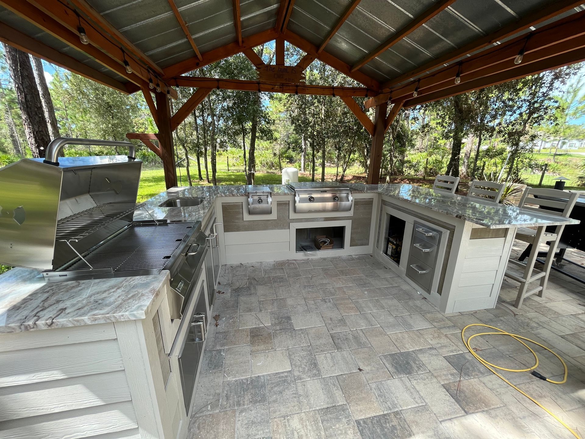 Outdoor kitchen with grill, counters, cabinets, and dining area under a wooden pavilion roof.