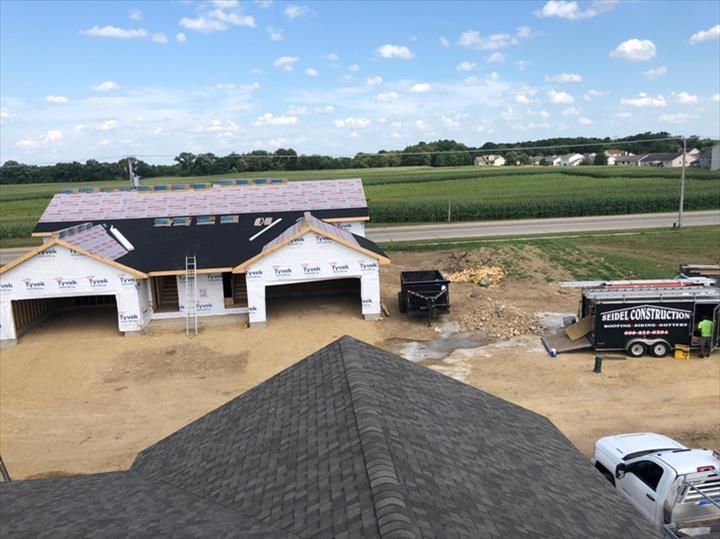 Construction site with a house under construction, trucks, dirt lot, and a partly built roof in the foreground