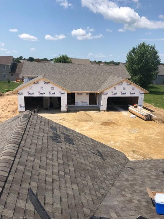 New house under construction with roof shingles, two garage openings, and dirt yard under a blue sky