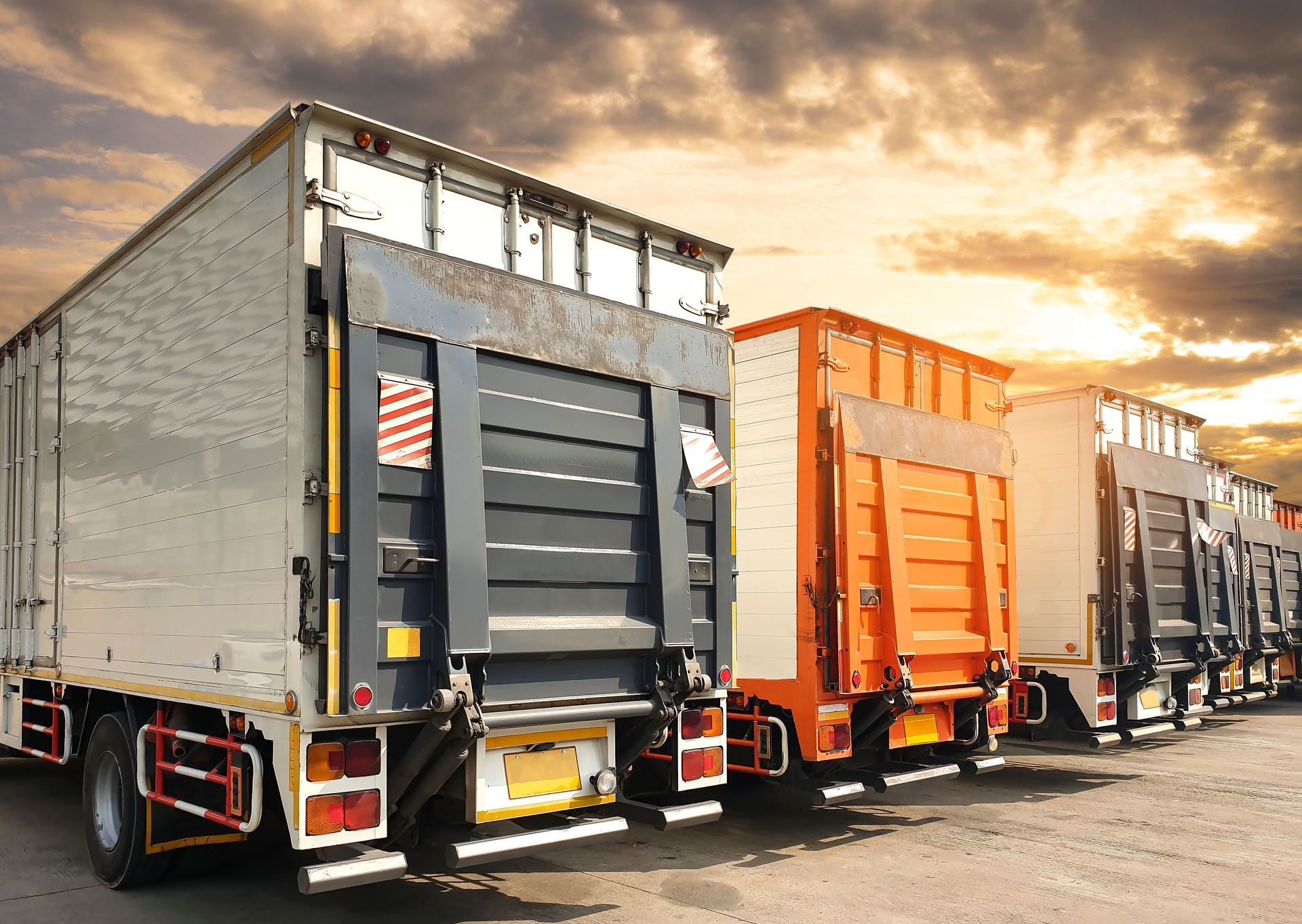 Delivery trucks parked outdoors, backs facing the viewer, against a sunset sky.