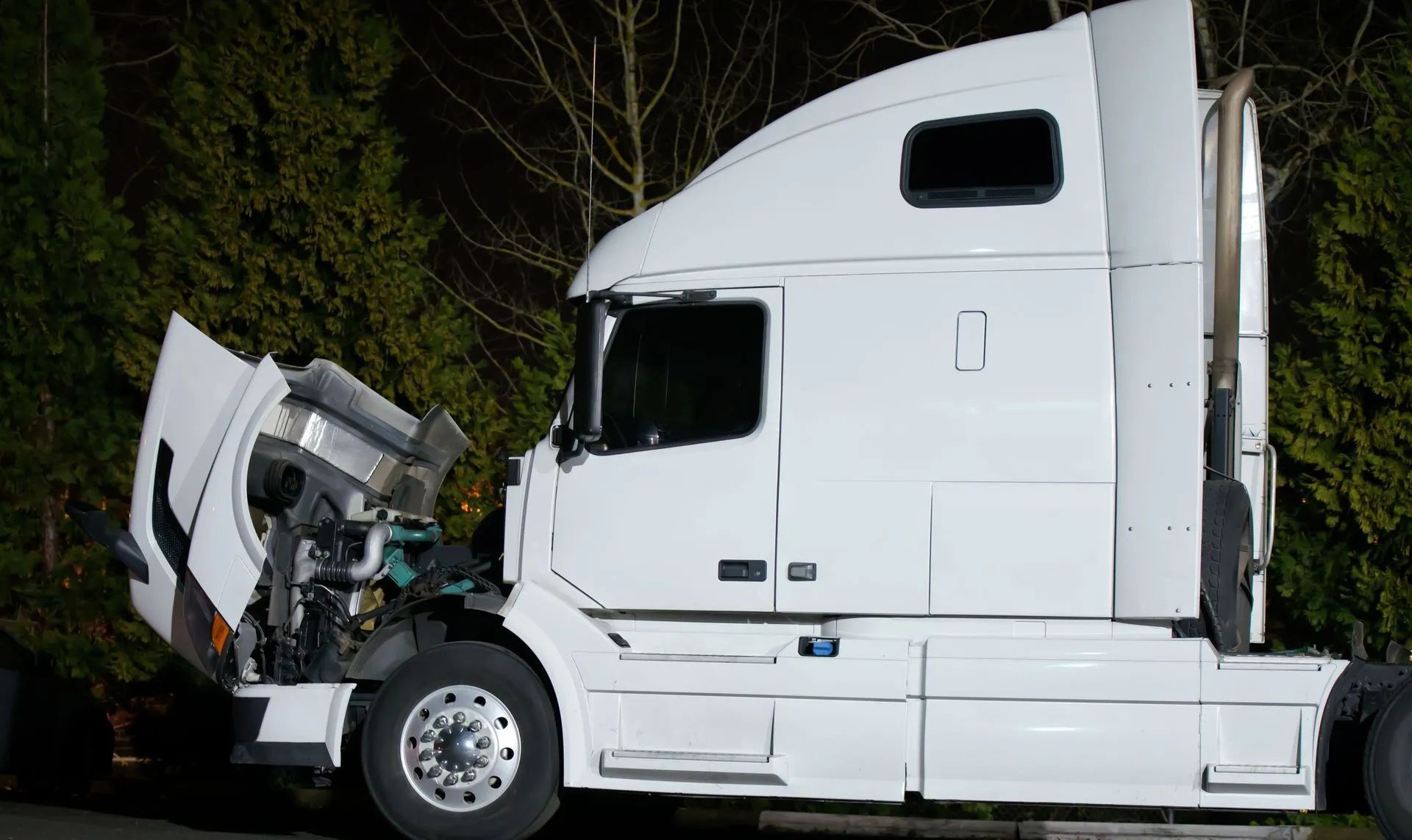 White semi-truck with open hood. The setting is outdoors at night, with green foliage in the background.