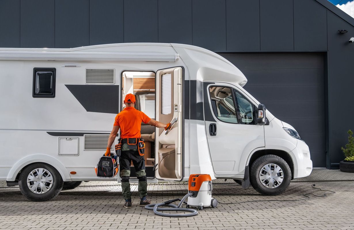 Man with tools enters white RV, vacuum cleaner at side, parked near a garage.