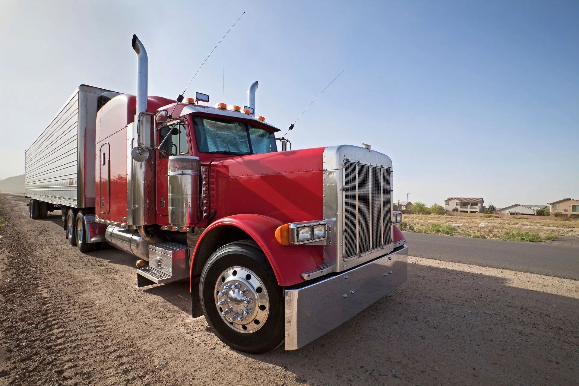 Red semi-truck parked on a dirt road next to a paved road on a sunny day.