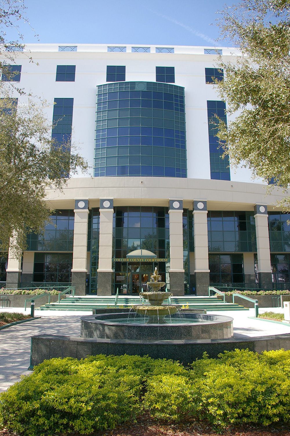 Modern building with blue glass facade, columns, and a fountain in front, framed by trees