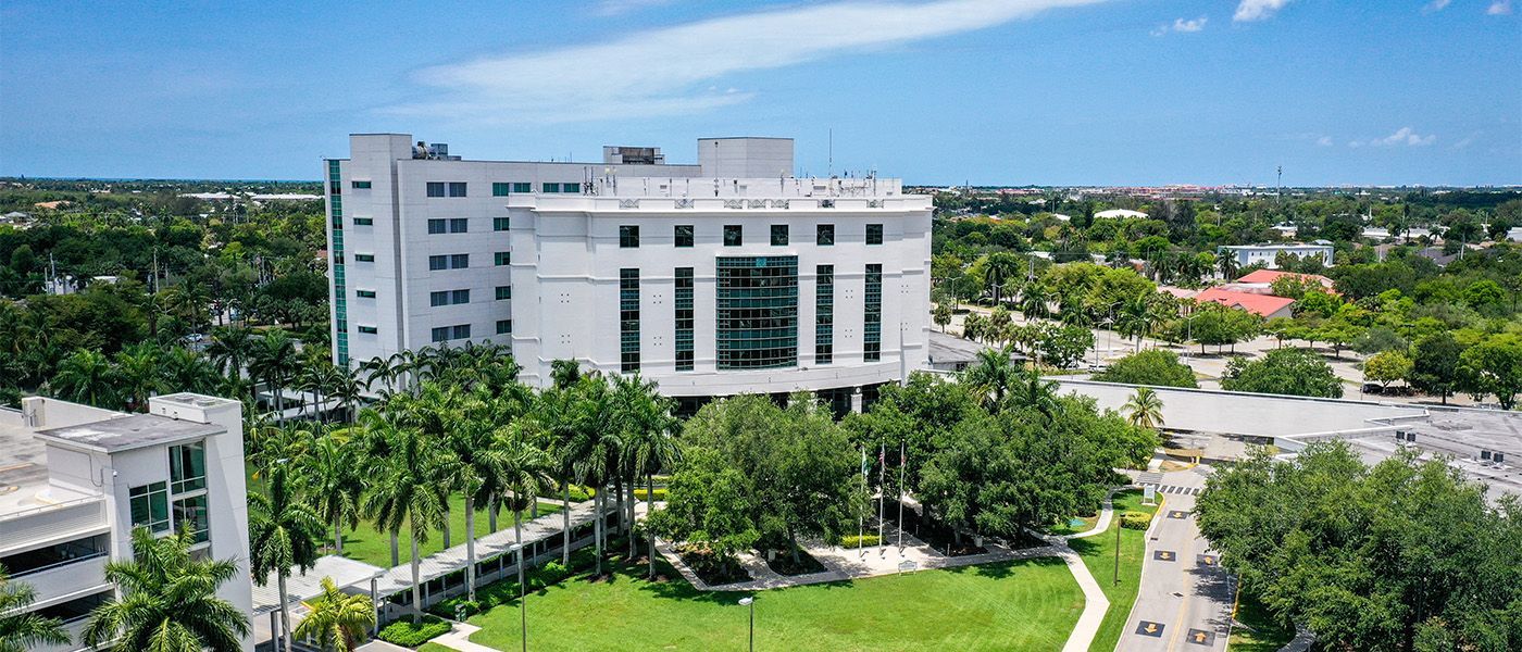 Aerial view of a white multi-story building surrounded by palm trees and green lawns under a blue sky